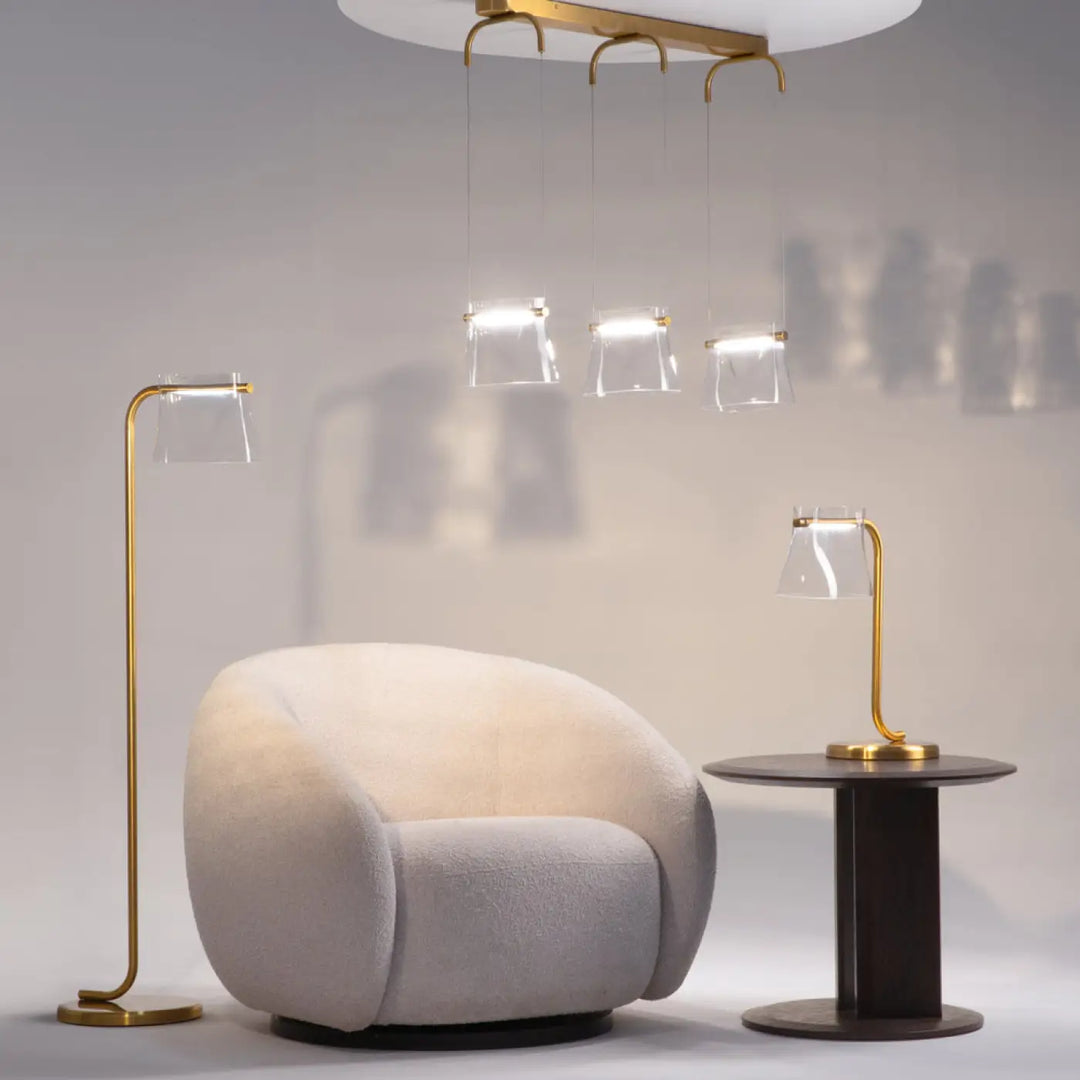 Fauteuil beige crème en tissu doux à design moderne arrondi présenté avec une lampe à poser en verre transparent aux lignes fluides