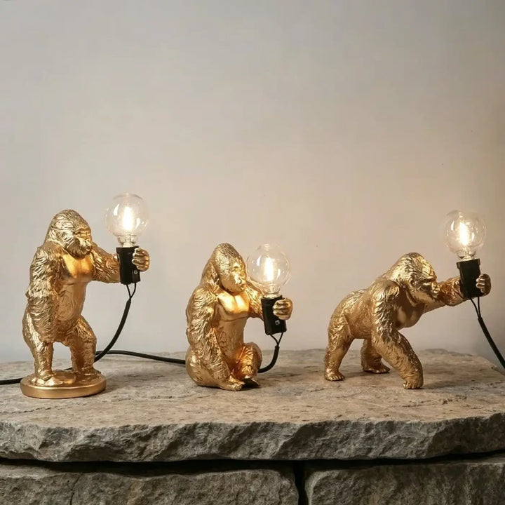 Lampe de table gorenzo avec trois lampes artistiques gorille dorées tenant des ampoules allumées sur pierre